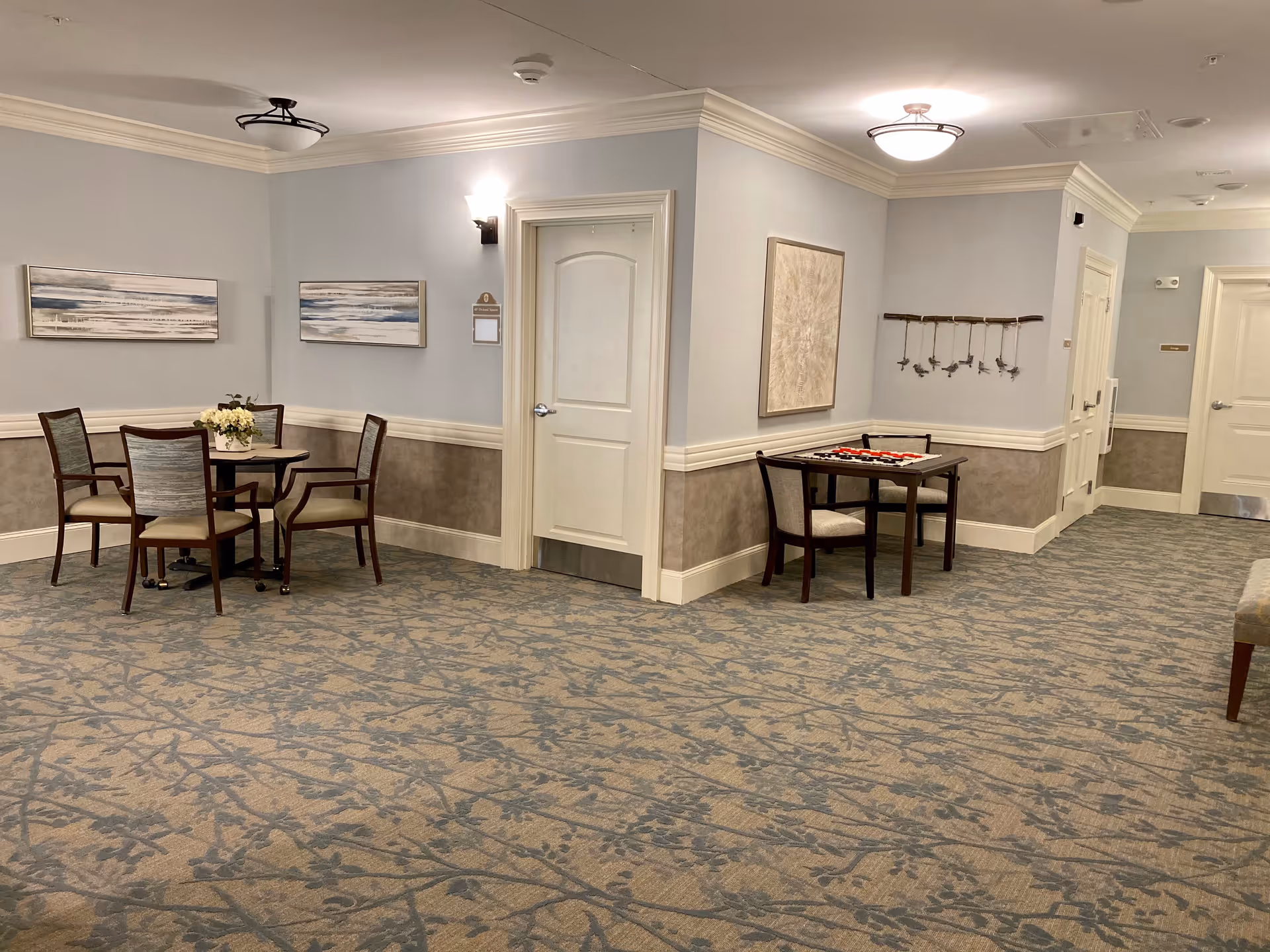 Interior view of a senior living facility common area with light blue walls and patterned carpet. There are two small tables with chairs; one table has a flower arrangement and the other has a board game set up. Several doors and framed artwork are visible on the walls.