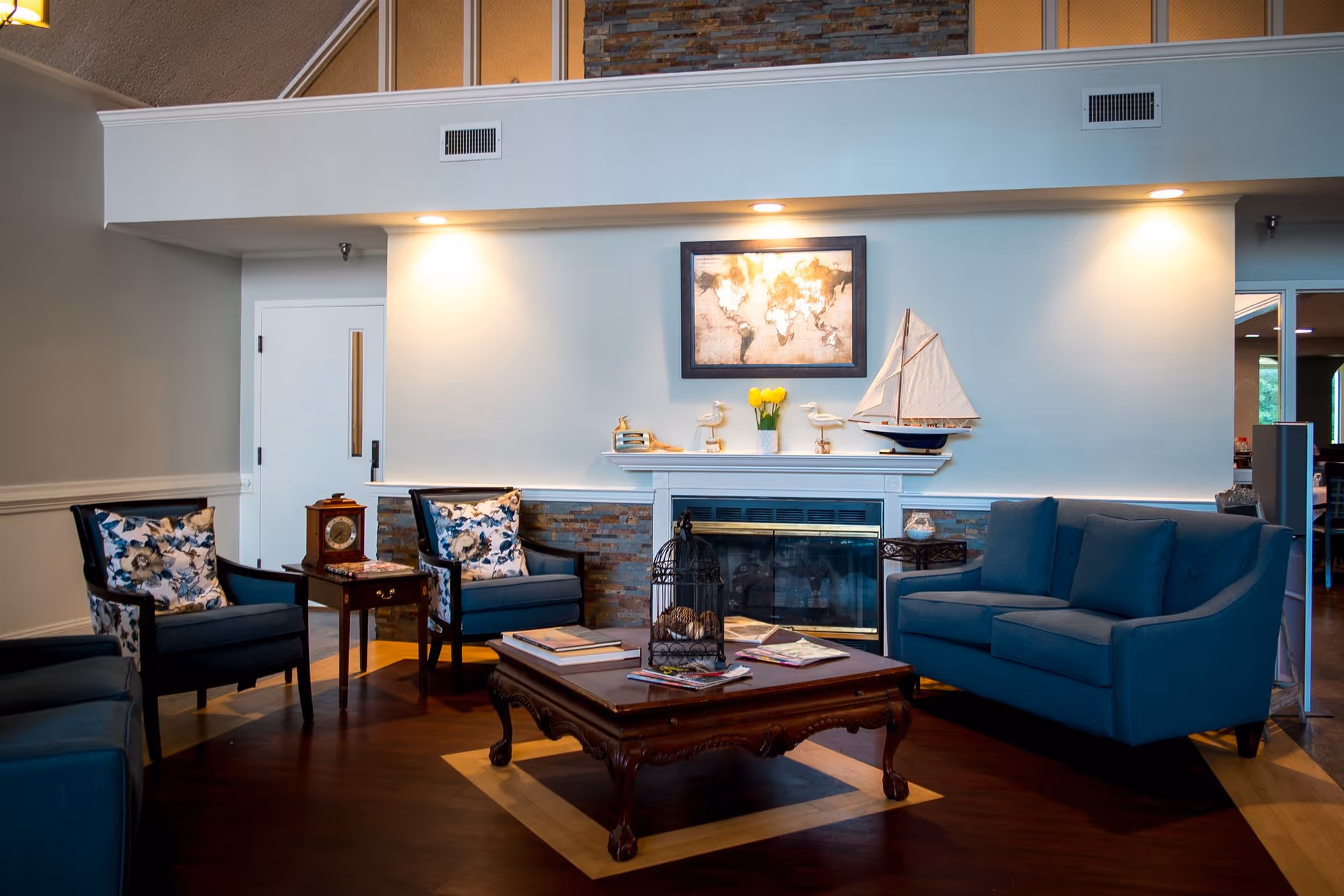A cozy living room with blue upholstered chairs and a sofa arranged around a wooden coffee table. The coffee table holds books and a decorative birdcage. The room features a fireplace with a white mantle decorated with a model sailboat, small sculptures, and a vase of yellow tulips. Above the fireplace hangs a framed world map. The walls are light-colored with recessed lighting above the fireplace, and the floor is dark wood with a lighter wood inlay.