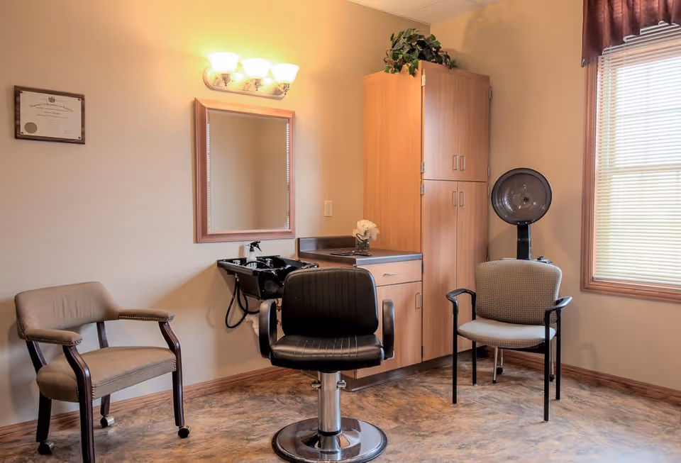 A small salon room with a black salon chair in front of a mirror and sink, two additional chairs, a wooden cabinet, a hair dryer, and a window with blinds and a curtain.