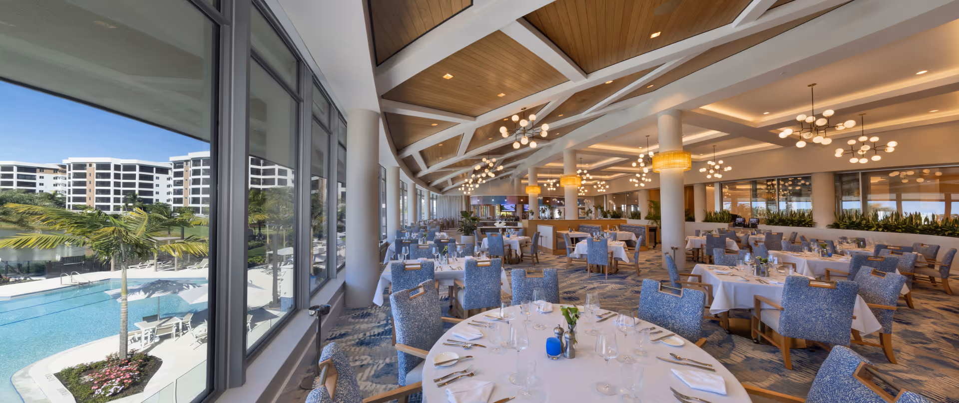 Spacious dining room with round tables covered in white tablecloths, set with glassware and silverware. Blue upholstered chairs surround the tables. Large windows on one side offer a view of a pool area with palm trees and multi-story buildings in the background. The ceiling features wooden panels and modern chandeliers providing warm lighting.