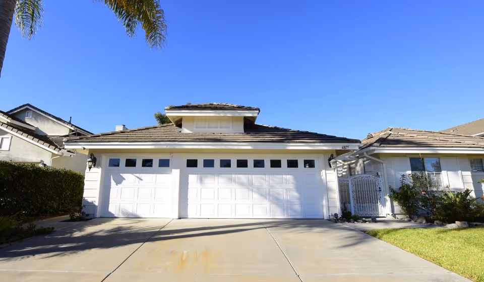 Exterior view of a residential building with a large white garage door, a driveway, and a small gated entrance to the right. The sky is clear and blue, and there are palm trees and shrubs around the property.