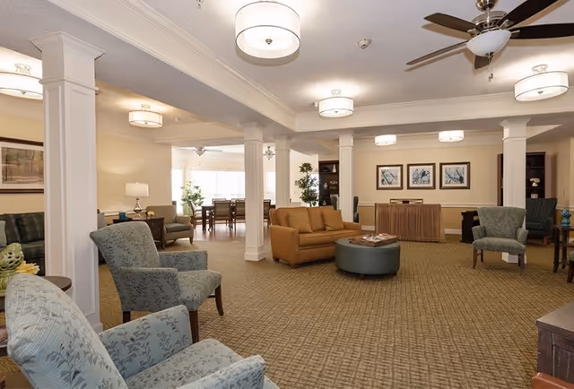 A spacious assisted living facility common area with multiple upholstered armchairs and a sofa arranged around a round ottoman. The room features beige walls, carpeted floors, white columns, ceiling fans, and modern ceiling lights. In the background, there is a dining area with tables and chairs near large windows letting in natural light. The room is decorated with framed artwork and plants.