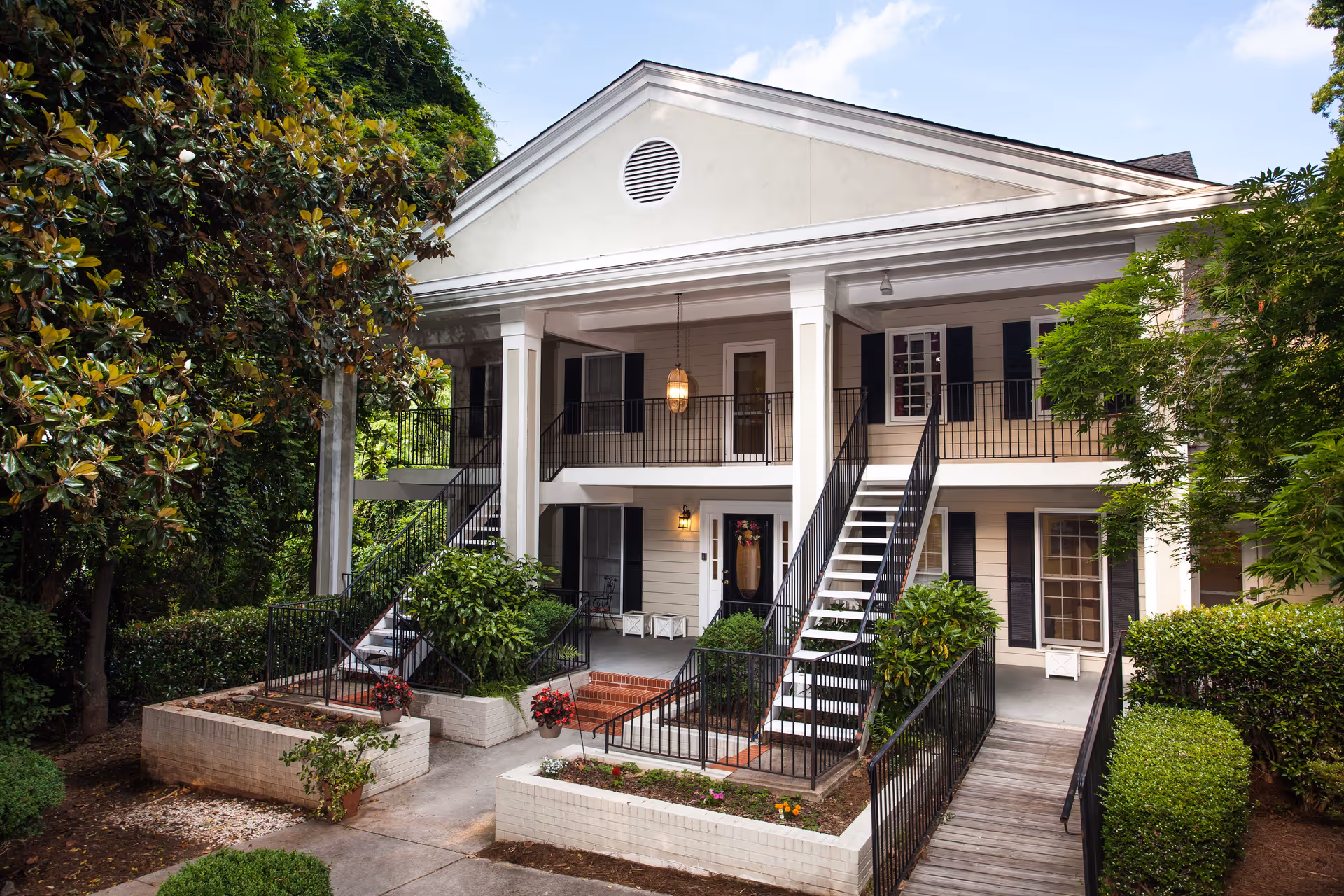 Exterior view of a two-story senior living facility building with white columns, black railings, and stairs leading to the upper floor. The entrance is surrounded by greenery, including bushes, trees, and flower beds. A hanging light fixture is visible on the upper porch.