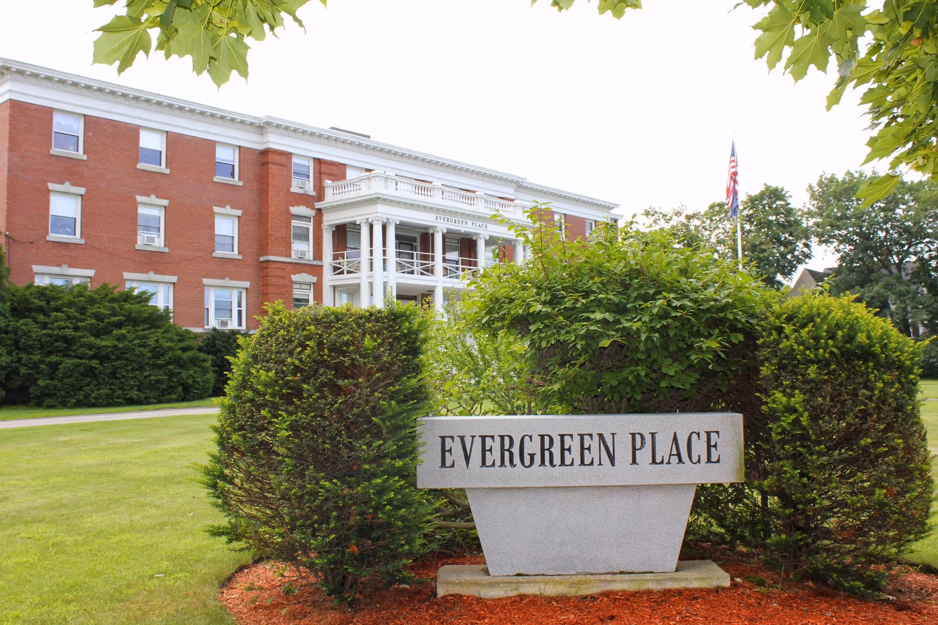Stone sign reading 'EVERGREEN PLACE' on a lawn in front of a red-brick, columned building.