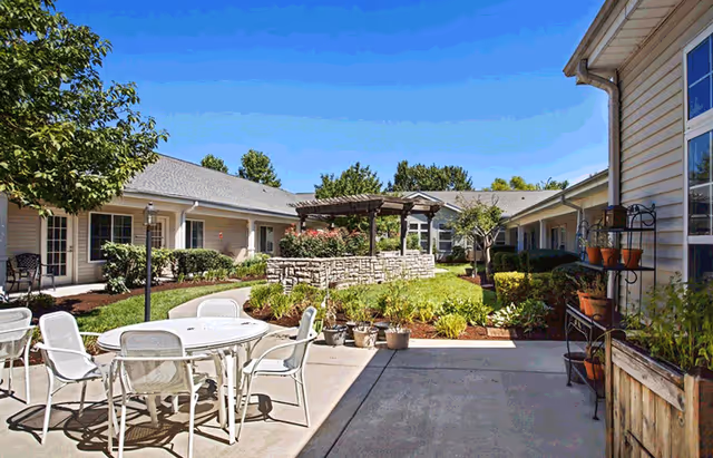 Outdoor courtyard area at American House Lebanon featuring a round white table with six white chairs on a concrete patio. Surrounding the patio are garden beds with various plants and flowers, a stone raised planter with a wooden pergola, and single-story building wings with windows and doors. The sky is clear and blue.