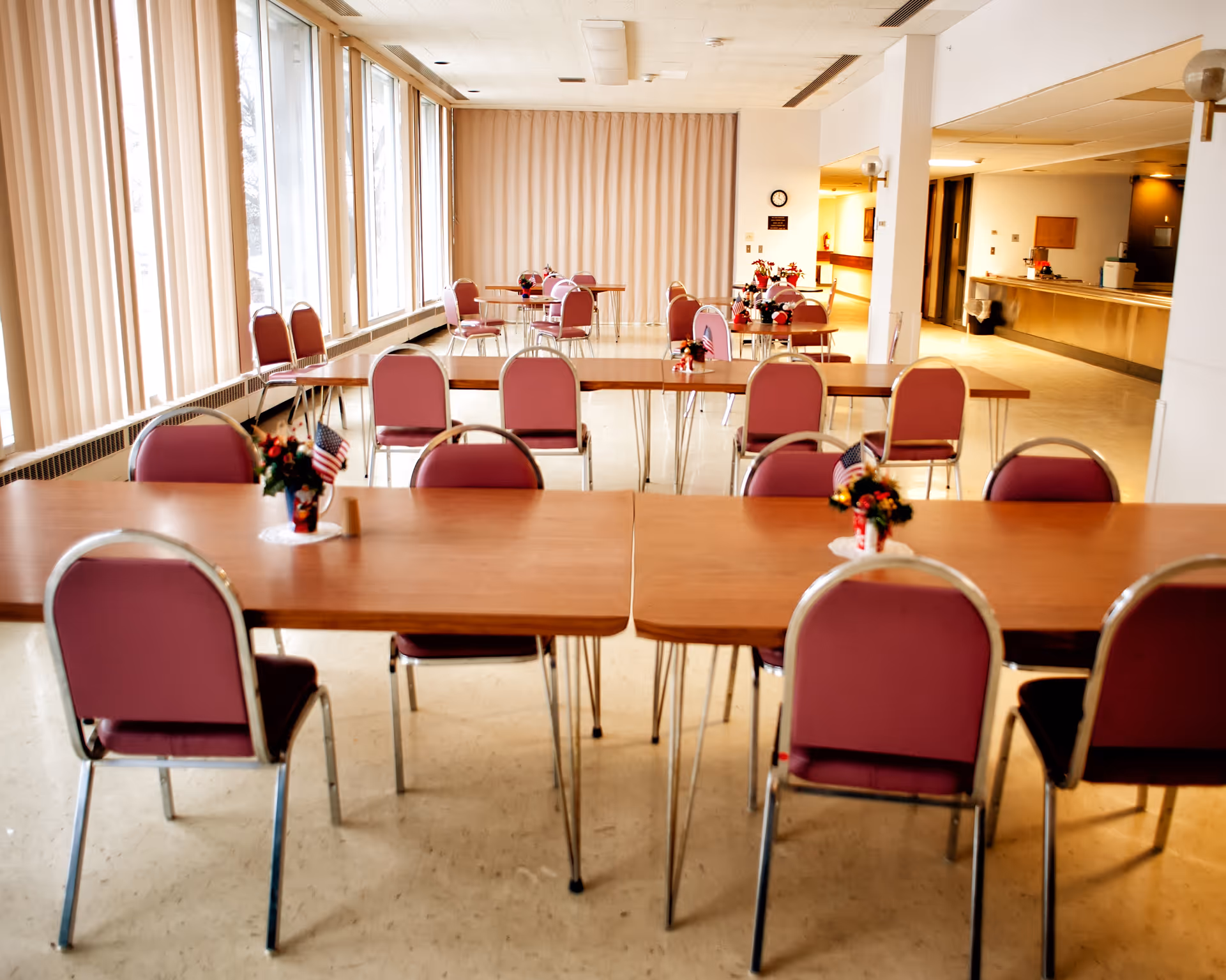 Communal dining room with rows of wooden tables and pink chairs, small floral centerpieces with American flags, and large windows with vertical blinds.