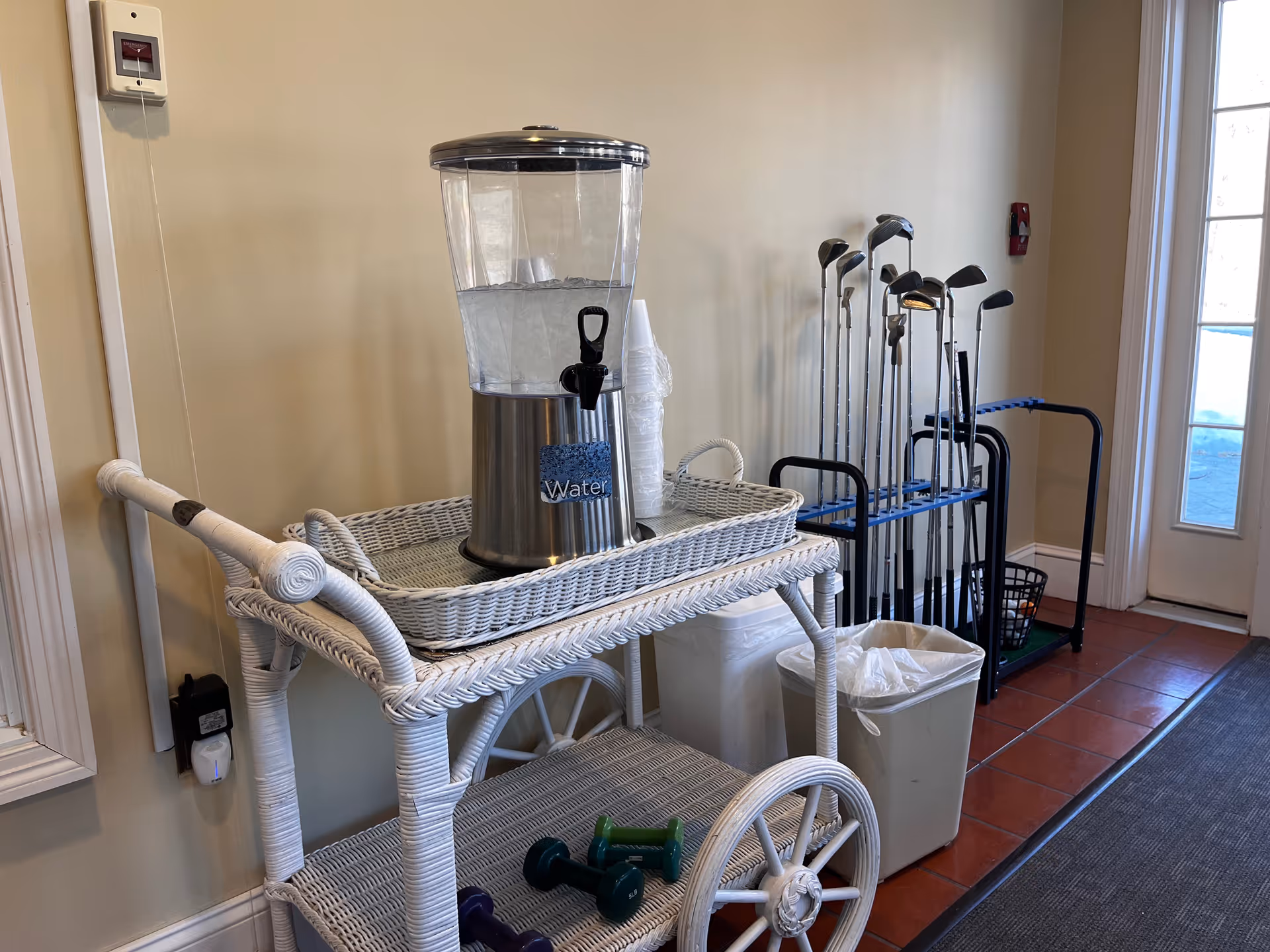 A white wicker cart with a large water dispenser labeled 'Water' and a stack of plastic cups on top. Below the cart are small hand weights. In the background, there is a rack holding several golf clubs next to a trash can and a door with glass panels.