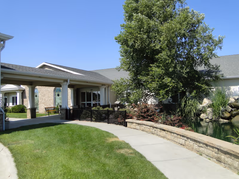 Covered entrance and walkway leading to the front of a single-story senior living building with landscaping, a pond, and trees.