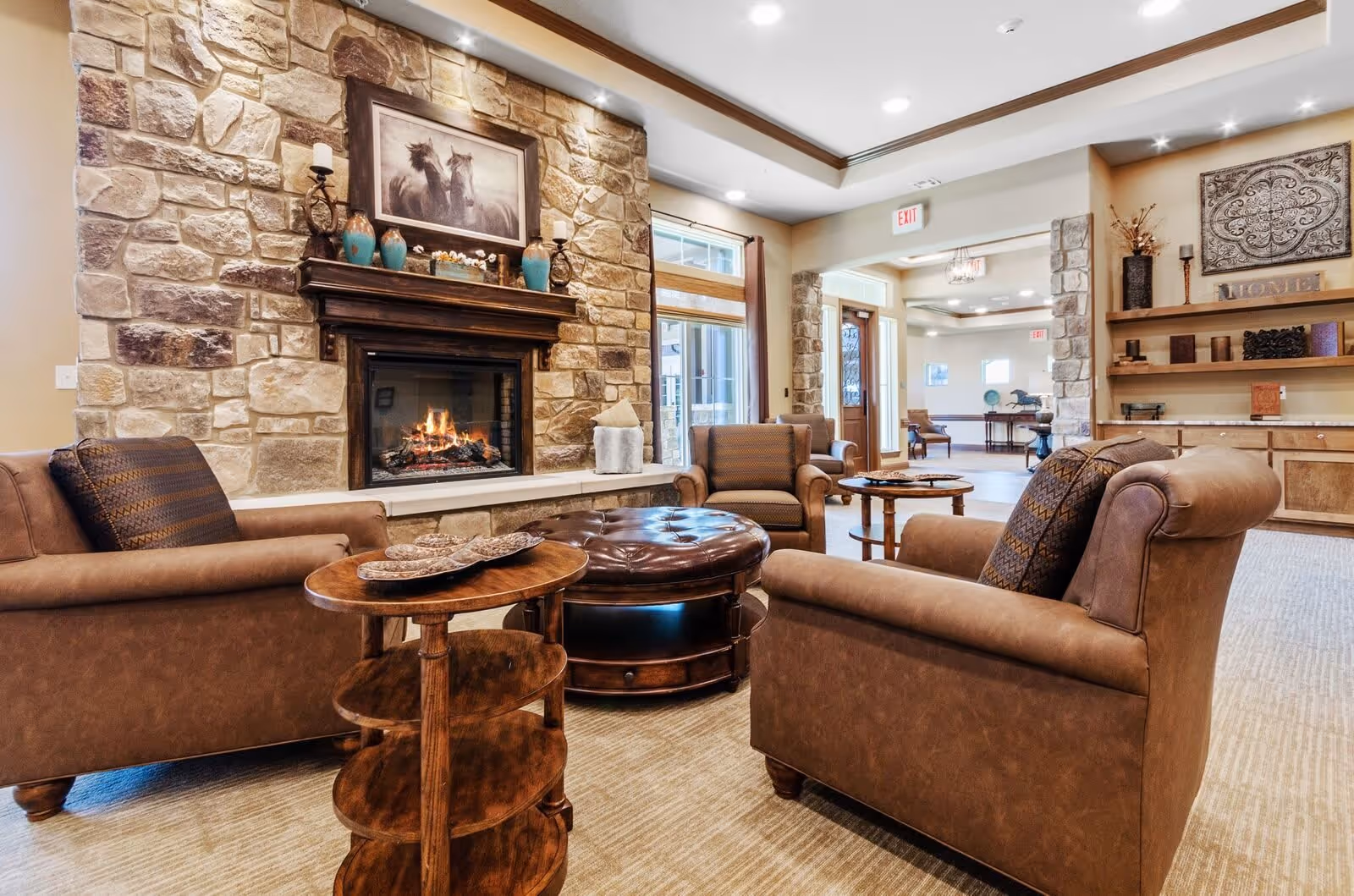 A cozy living room area with a stone fireplace featuring a painting of horses above the mantle. The room has brown leather armchairs with patterned cushions arranged around a round leather ottoman and a wooden side table. The walls are beige with stone accents, and there are shelves with decorative items on the right side. The space is well-lit with recessed lighting and natural light from windows and doors.