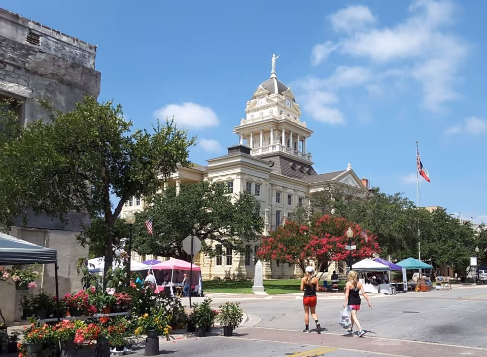 Historic courthouse-style building with a domed tower, trees and market tents in front and two people crossing the street.
