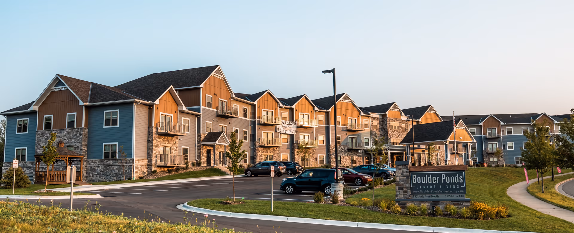 Exterior view of Boulder Ponds Senior Living facility showing a large multi-story building with a combination of blue and brown siding and stone accents. The building features multiple balconies and a parking lot with several cars parked. There is a sign in the foreground that reads 'Boulder Ponds Senior Living' along with the website and phone number. The scene is set during daylight with a clear sky.