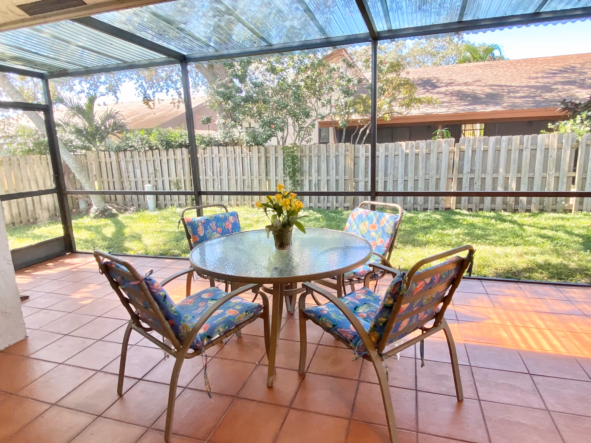 A screened-in patio with a round glass table and four metal chairs with colorful floral cushions. A small potted plant with yellow flowers is on the table. The patio overlooks a grassy backyard enclosed by a wooden fence, with trees and neighboring houses visible in the background.