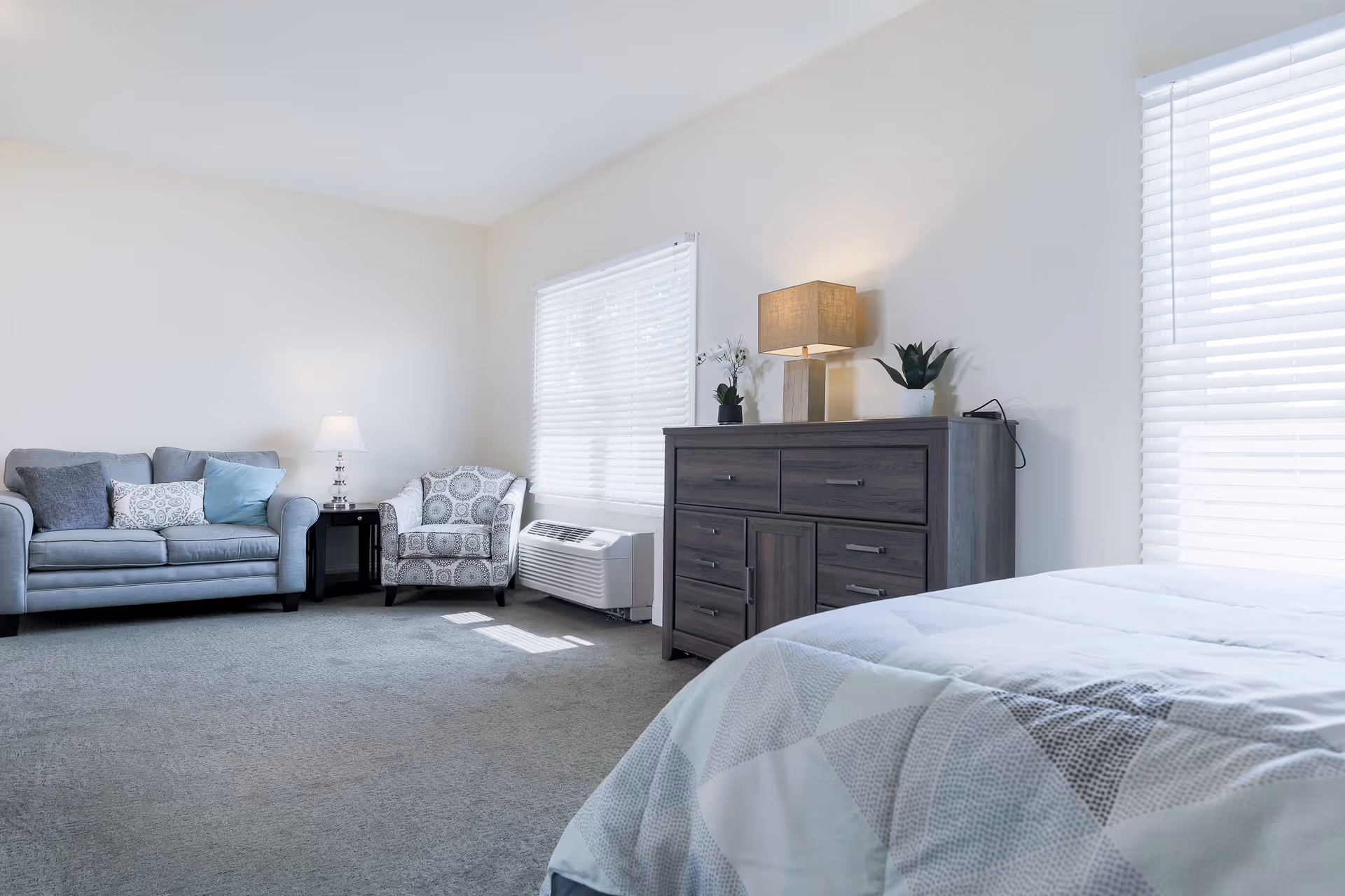 A bright and clean bedroom in an assisted living facility featuring a bed with a patterned quilt, a wooden dresser with a lamp and plants on top, a window with blinds, a gray sofa with cushions, a patterned armchair, and a small side table with a lamp.