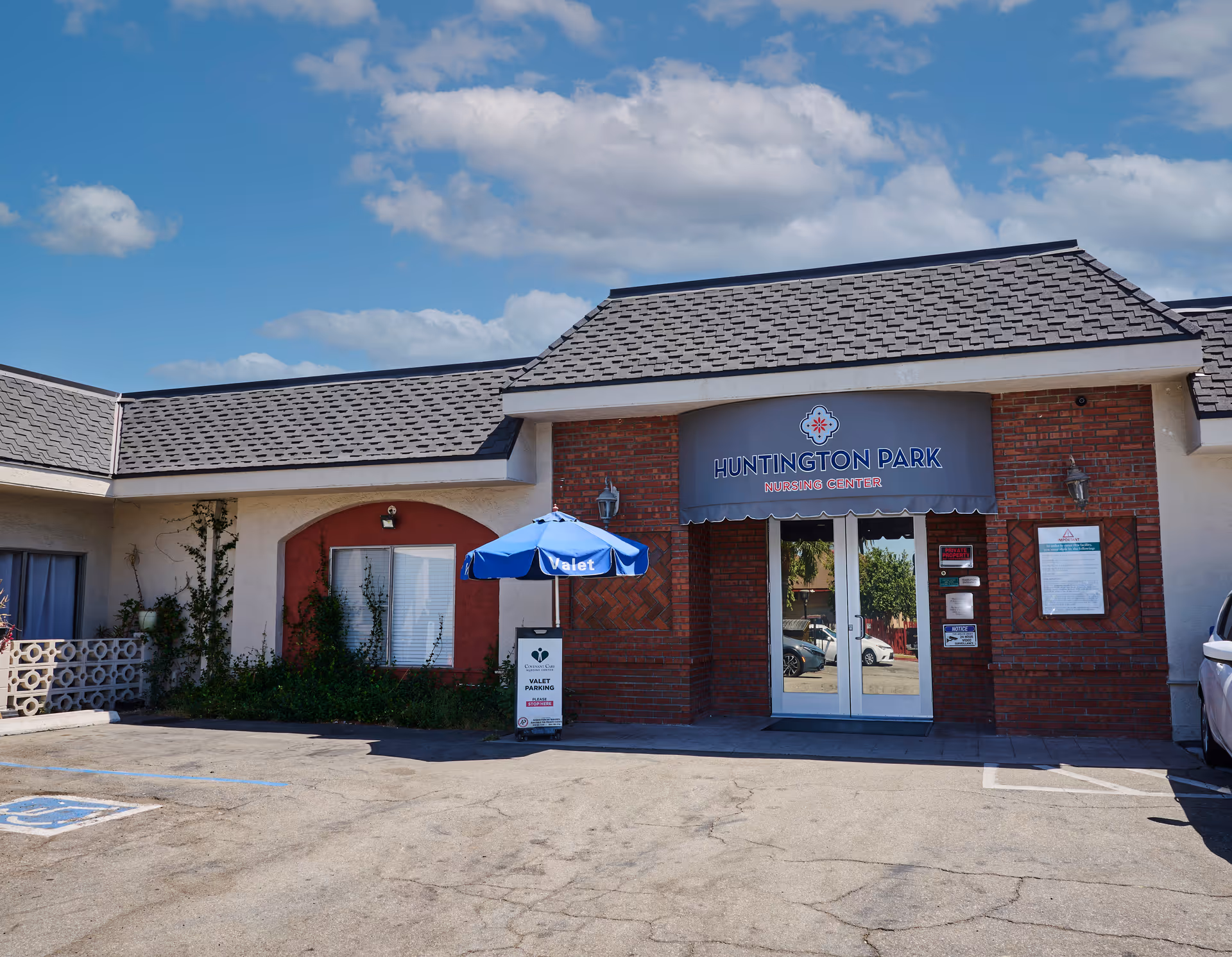 Exterior front entrance of Huntington Park Nursing Center with a gray shingled roof, brick and stucco walls, a blue valet umbrella, and a sign indicating valet parking. The sky is blue with scattered clouds.