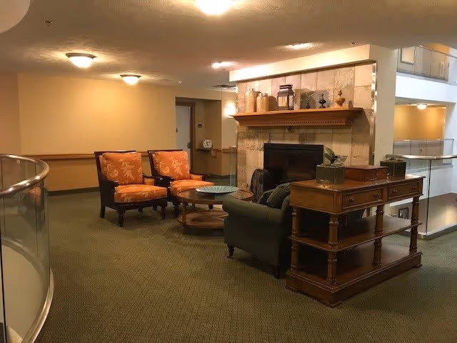 A cozy seating area in a retirement facility featuring two orange upholstered armchairs, a green sofa, a round glass coffee table, and a wooden side table with drawers. The room has a fireplace with decorative items on the mantel and soft lighting from ceiling fixtures. The carpet is green and the walls are painted beige with a wooden chair rail.