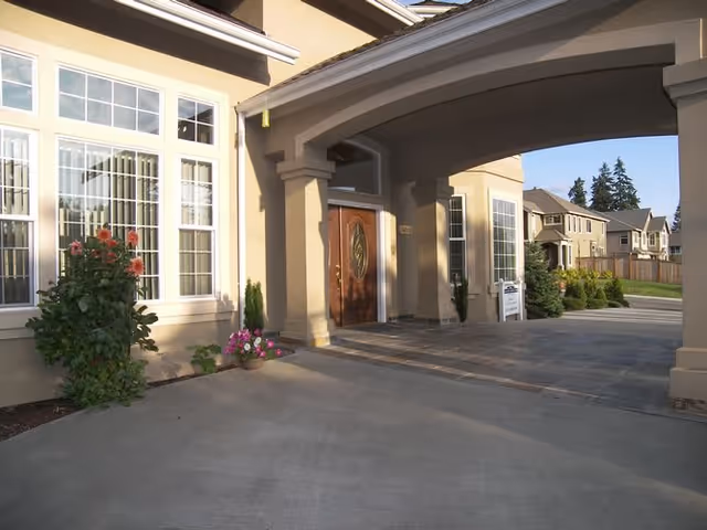 Covered driveway and main entrance to a residential building with large windows, double wooden doors, and potted plants.