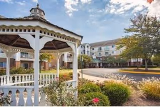 Outdoor view of a senior living facility featuring a white gazebo in the foreground, landscaped bushes and flowers, a paved driveway, and a multi-story building with balconies in the background under a partly cloudy sky.