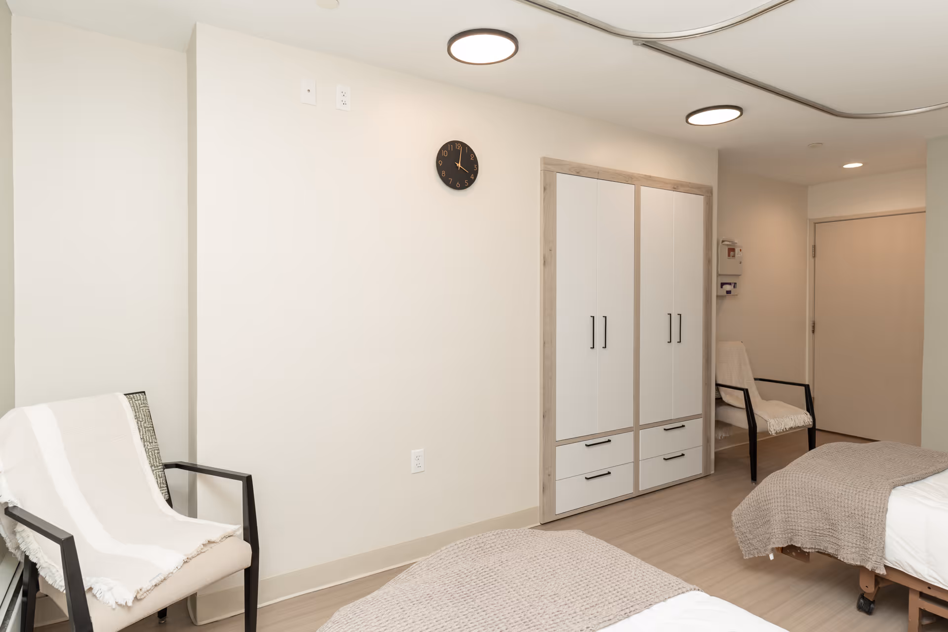 A clean and simple bedroom in a senior living facility with two beds covered in beige blankets, two chairs with white throws, a white wardrobe with wooden trim, a wall clock, and soft overhead lighting.