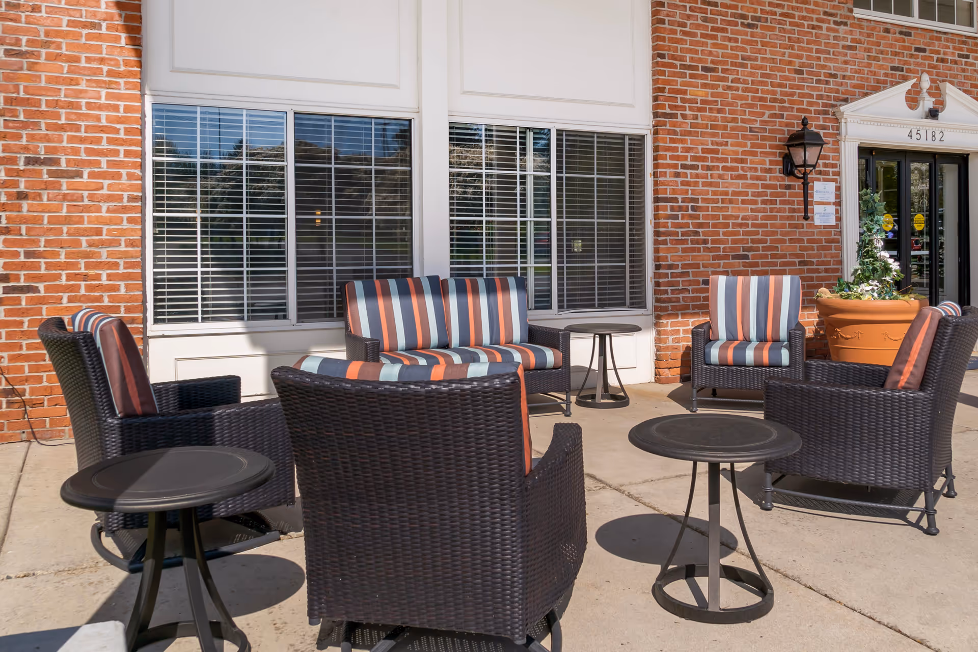 Outdoor seating area with black wicker chairs and a loveseat featuring striped cushions in shades of orange, blue, and gray. The seating is arranged around small round black tables on a concrete patio in front of a brick building with large windows and a door marked 45182.
