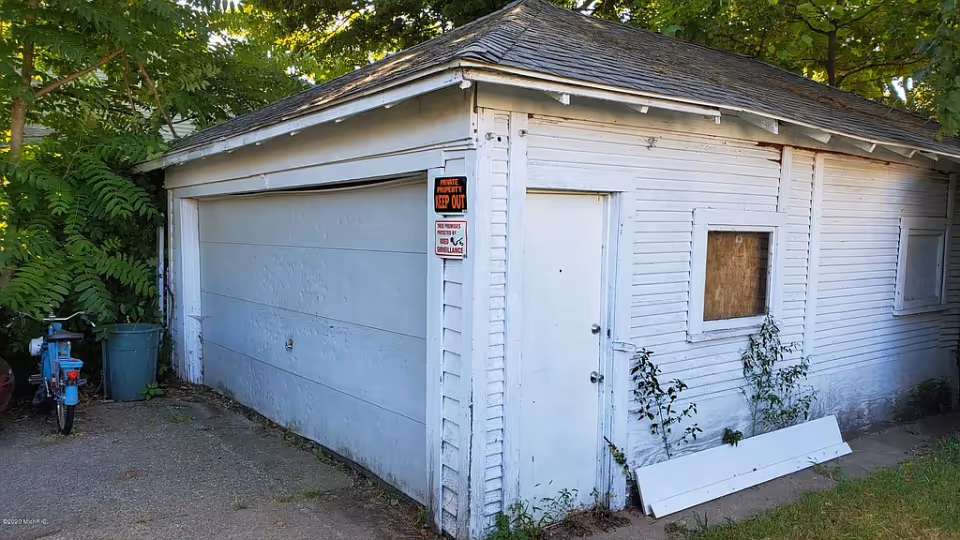 White detached garage/outbuilding with a closed garage door, a side entry door, a boarded window, and plants growing around it.