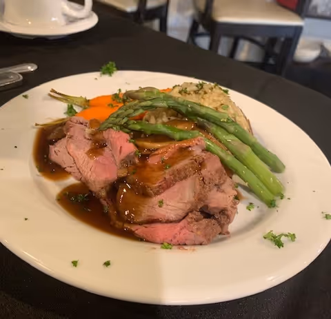 A plate of food featuring slices of medium-rare roast beef topped with brown gravy, served with steamed asparagus, cooked carrots, and a portion of rice pilaf, garnished with chopped parsley. The plate is on a black tablecloth with a white coffee cup and saucer in the background.