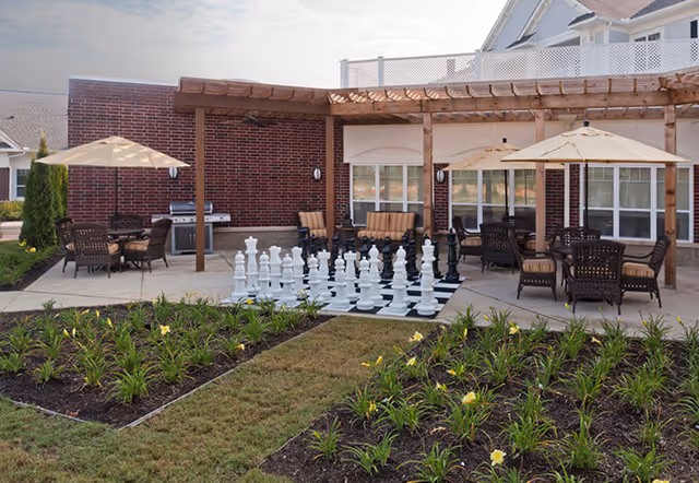 Outdoor patio area at Irene Woods Assisted Living featuring a large chessboard with oversized chess pieces, several seating areas with tables and umbrellas, a pergola, and a brick building in the background surrounded by landscaped flower beds.