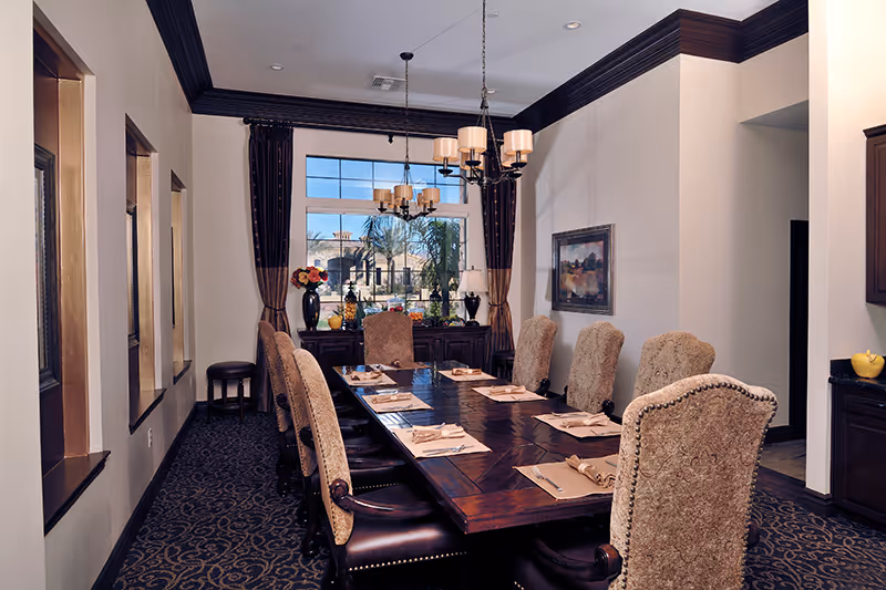 A formal dining room with a long wooden table set for eight people. The chairs are upholstered with a patterned fabric and have dark wooden legs. The table is set with placemats, napkins, and silverware. A chandelier with multiple lights hangs above the table. There is a large window with dark curtains at the far end of the room, showing an outdoor view with palm trees and a building. The walls are light-colored with dark crown molding, and there is a framed painting on one wall.