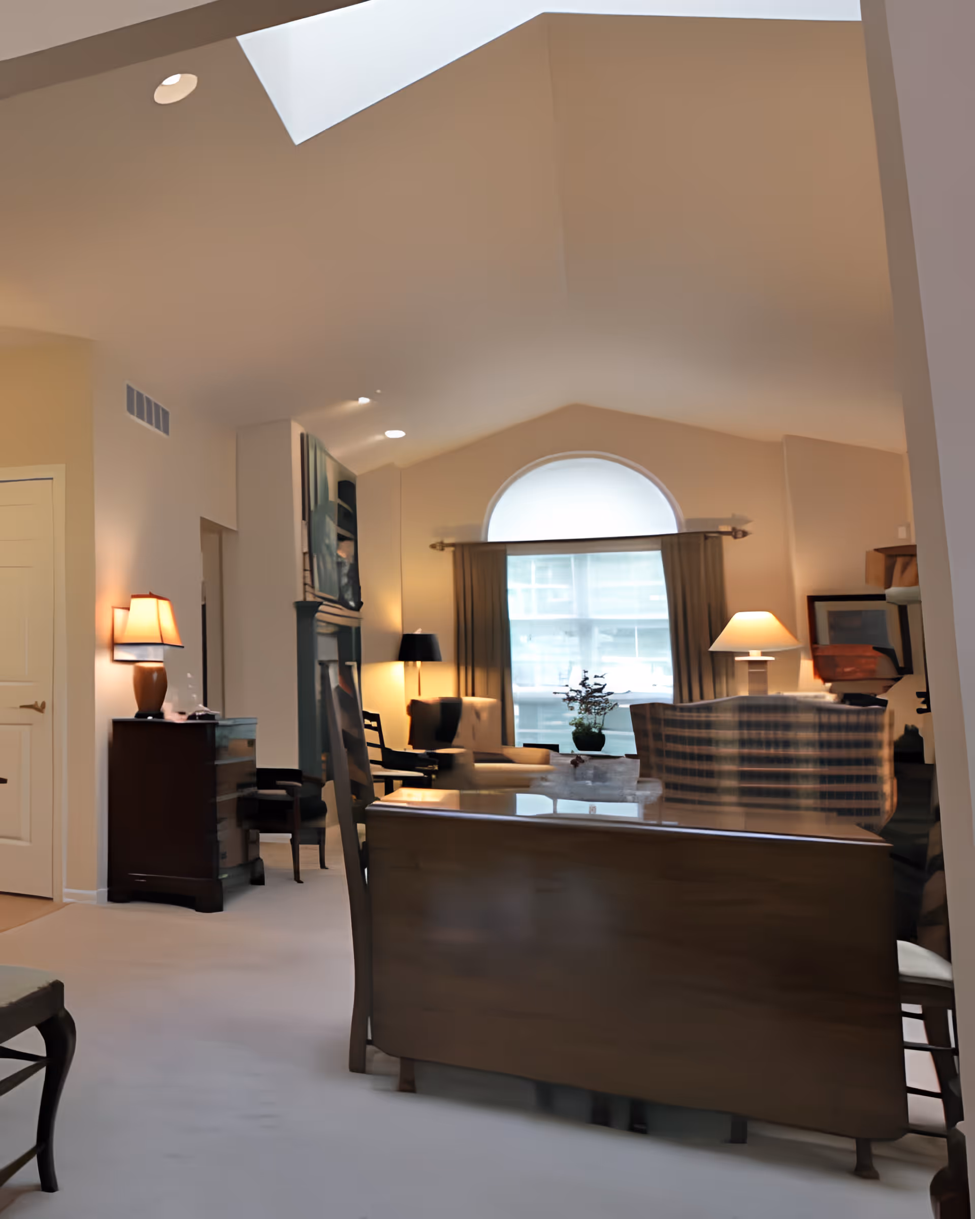 A cozy living room with a vaulted ceiling and a skylight. The room features a large arched window with curtains, a variety of lamps providing warm lighting, a wooden dining table with chairs, and comfortable seating including an armchair and a plaid sofa. There is a small plant on a table near the window and artwork on the walls.