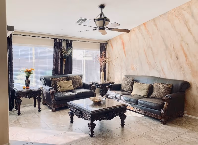 A bright living room with two dark leather sofas adorned with decorative pillows, a matching ornate wooden coffee table, and two side tables. The room has large windows with white blinds and dark curtains, a ceiling fan with light, and a wall with a marble-like texture.