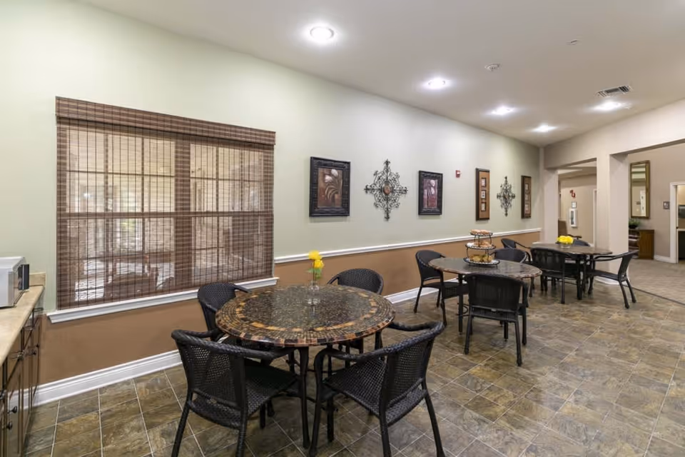 Interior view of a dining area in an assisted living facility with three round tables, each surrounded by black wicker chairs. The tables have decorative items including a small vase with yellow flowers and a tiered tray. The walls are painted in two tones with framed artwork and decorative metal wall hangings. There is a window with brown blinds and a countertop with a microwave on the left side. The floor is tiled and the ceiling has recessed lighting.