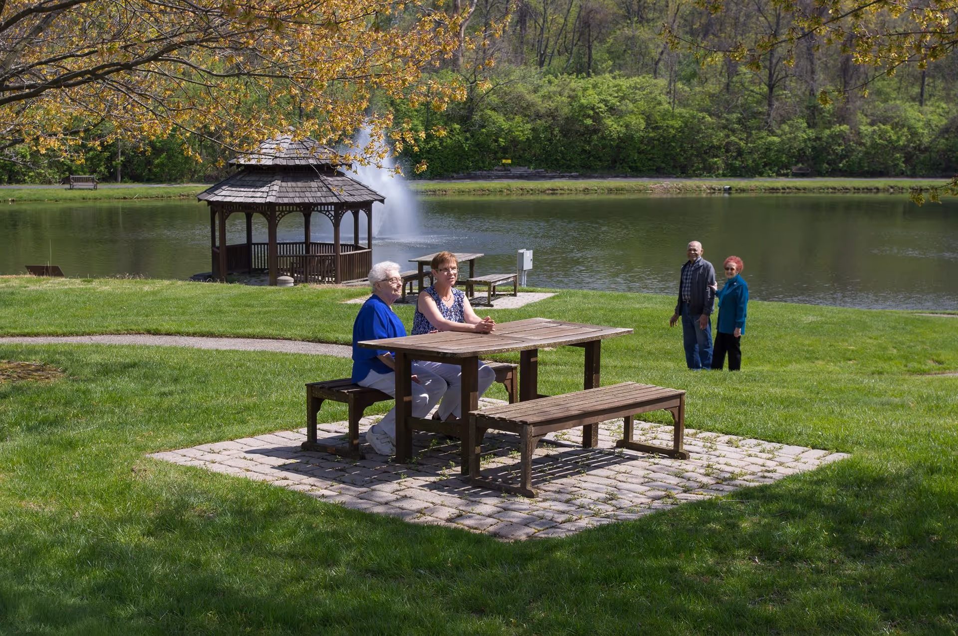 Two elderly women sitting at a wooden picnic table on a stone patio near a pond with a fountain and a gazebo in the background. Two other people are standing on the grass near the pond. The scene is set in a green, park-like outdoor area with trees and a walking path.