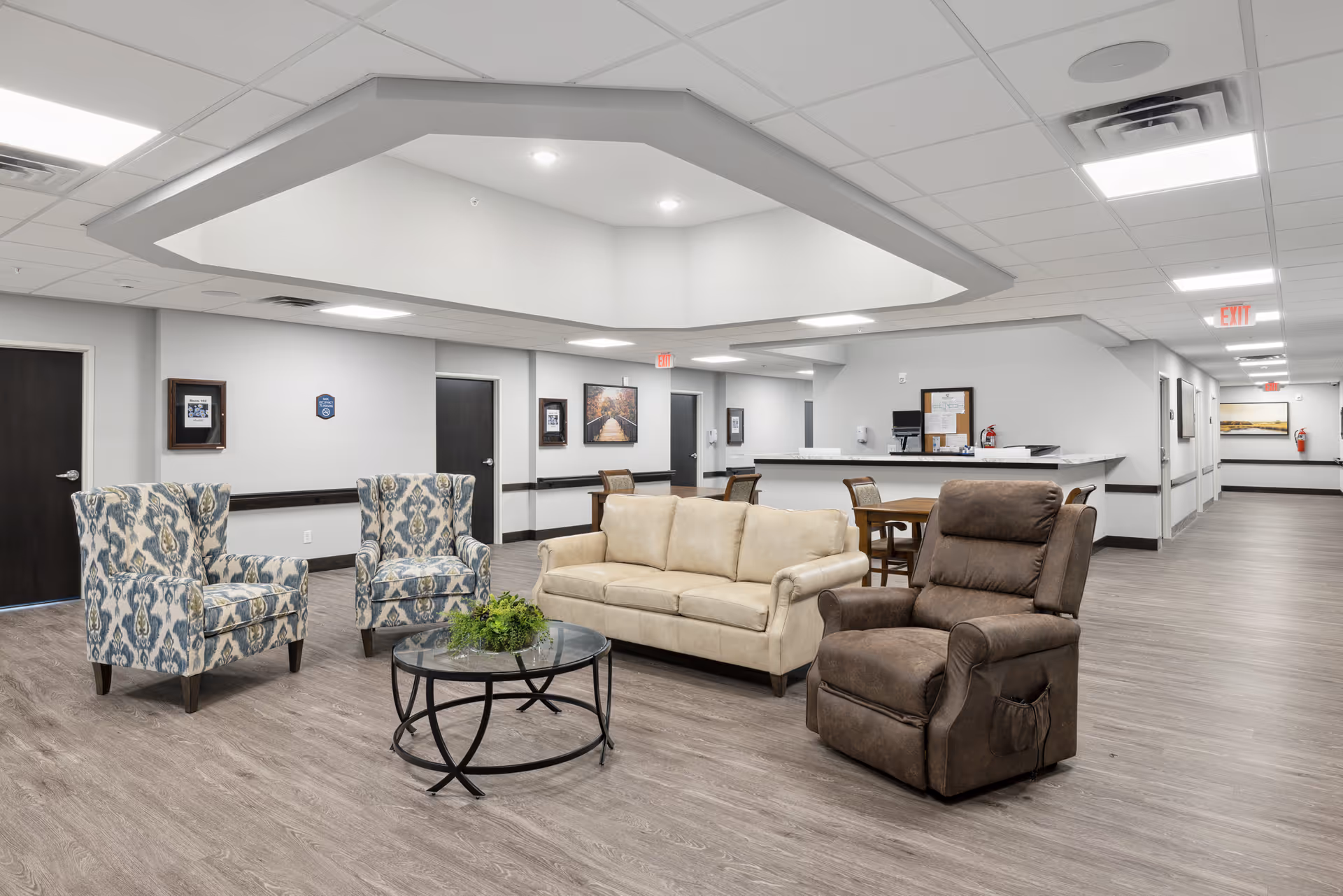 A spacious senior living facility common area with modern decor featuring two patterned armchairs, a beige sofa, and a brown recliner arranged around a round glass coffee table with a plant centerpiece. The room has light gray walls, wood-look flooring, recessed lighting, and a reception desk in the background with a bulletin board and computer. Several doors and framed pictures are visible along the walls.