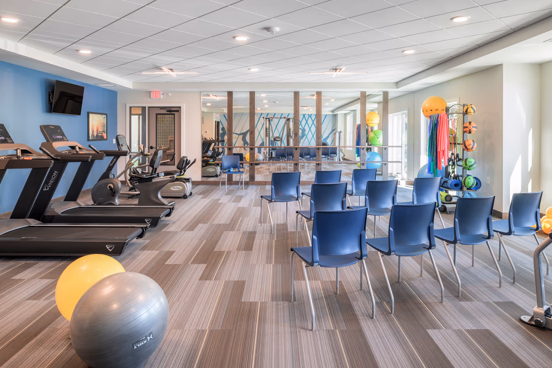 A fitness room with several treadmills and exercise bikes on the left side, a row of blue chairs arranged in the center, and various exercise balls and equipment on the right side. The room has a large mirror on the far wall and a blue accent wall with a mounted TV.