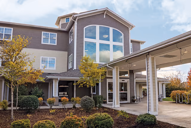 Exterior view of a multi-story senior living facility building with large windows, a covered entrance, and landscaped bushes and trees in front under a partly cloudy sky.