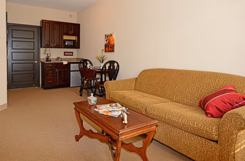 A cozy living area in a retirement center featuring a patterned beige sofa with a red pillow, a wooden coffee table with magazines and a small figurine, a small dining table with two chairs, and a kitchenette with dark wood cabinets, a microwave, and a mini refrigerator. The walls are light-colored and there is a closed dark door in the background.