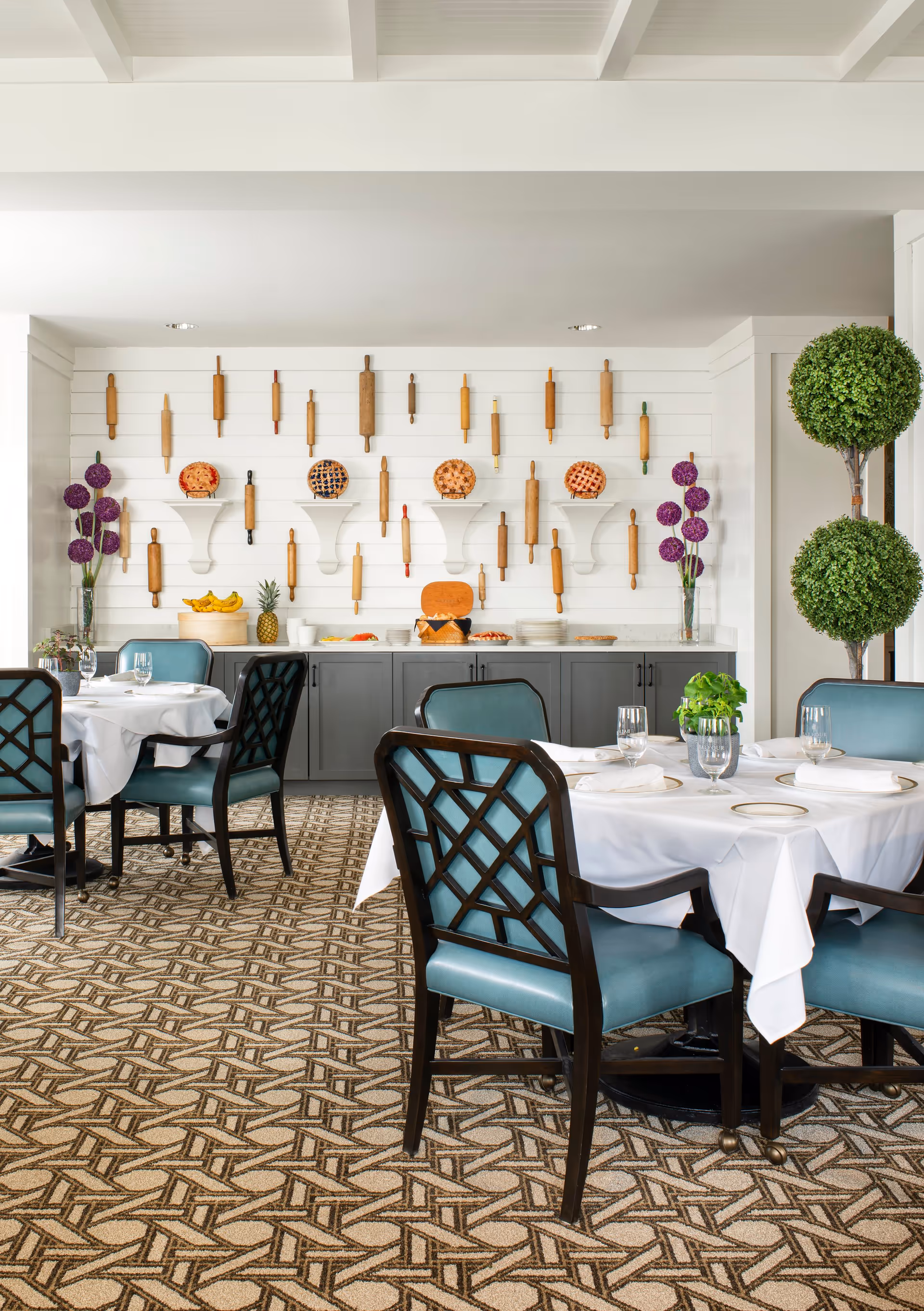 A bright dining room with tables covered in white tablecloths and set with plates, glasses, and napkins. The chairs have dark wooden frames with blue upholstery. The back wall features a decorative display of various rolling pins and several pies on white shelves. There are two tall vases with purple flowers and a double-tiered green topiary plant on the right side. The floor is carpeted with a geometric pattern.