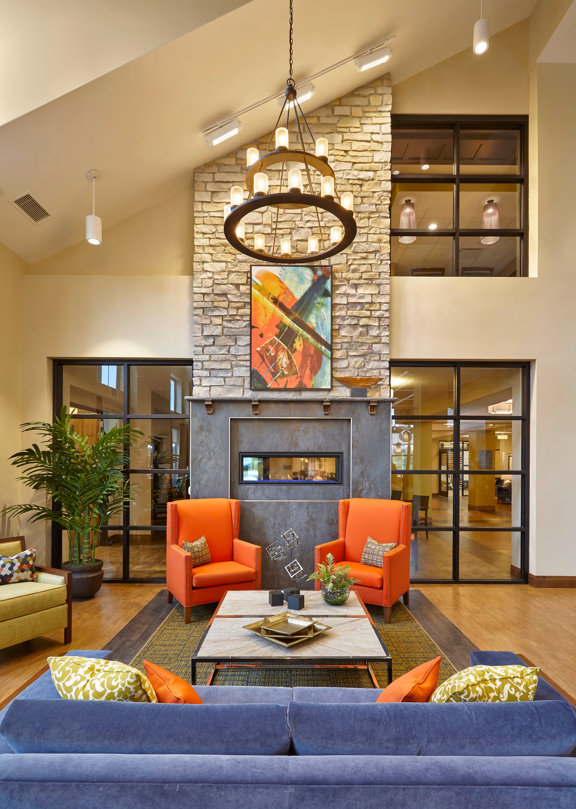 A cozy living room area with a stone fireplace and modern electric insert. Two bright orange armchairs with patterned pillows face a square coffee table decorated with geometric sculptures and a small plant. A blue sofa with colorful pillows is positioned in the foreground. The room features high ceilings with a large circular chandelier and track lighting. Large windows and glass doors reveal adjacent rooms and hallways.
