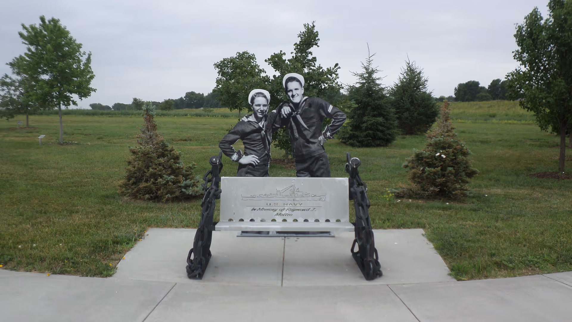 A memorial bench flanked by decorative anchors and life-size cutout figures of two sailors on a concrete path in a grassy park.