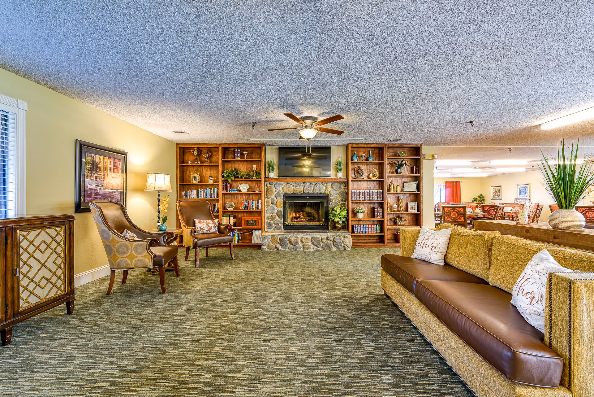 A cozy living room area in a senior living facility featuring a stone fireplace with a TV mounted above it, flanked by wooden bookshelves filled with books and decorative items. There are two upholstered armchairs with a floor lamp and framed artwork on the left side, and a long sofa with decorative pillows on the right. The room has carpeted flooring, a ceiling fan, and an open view into a dining area in the background.