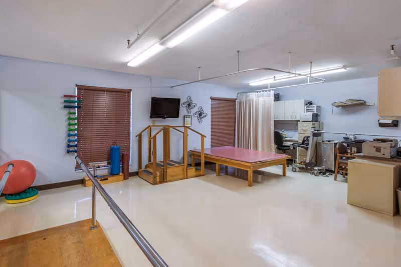 A rehabilitation room with parallel bars for walking exercises, a small set of stairs with handrails, a padded therapy table, exercise equipment including a large red exercise ball and foam rollers, and an office area with desks, chairs, and filing cabinets. The room has light-colored walls, two windows with brown blinds, and fluorescent ceiling lights.