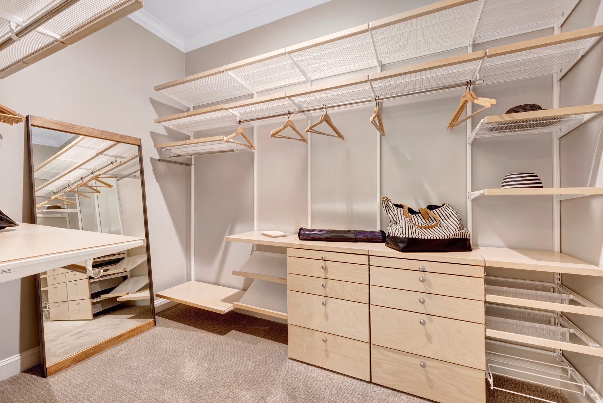 A spacious walk-in closet with multiple wooden shelves, drawers, and hanging rods with wooden hangers. There is a large floor mirror leaning against the wall and a striped bag and hat placed on the shelves.