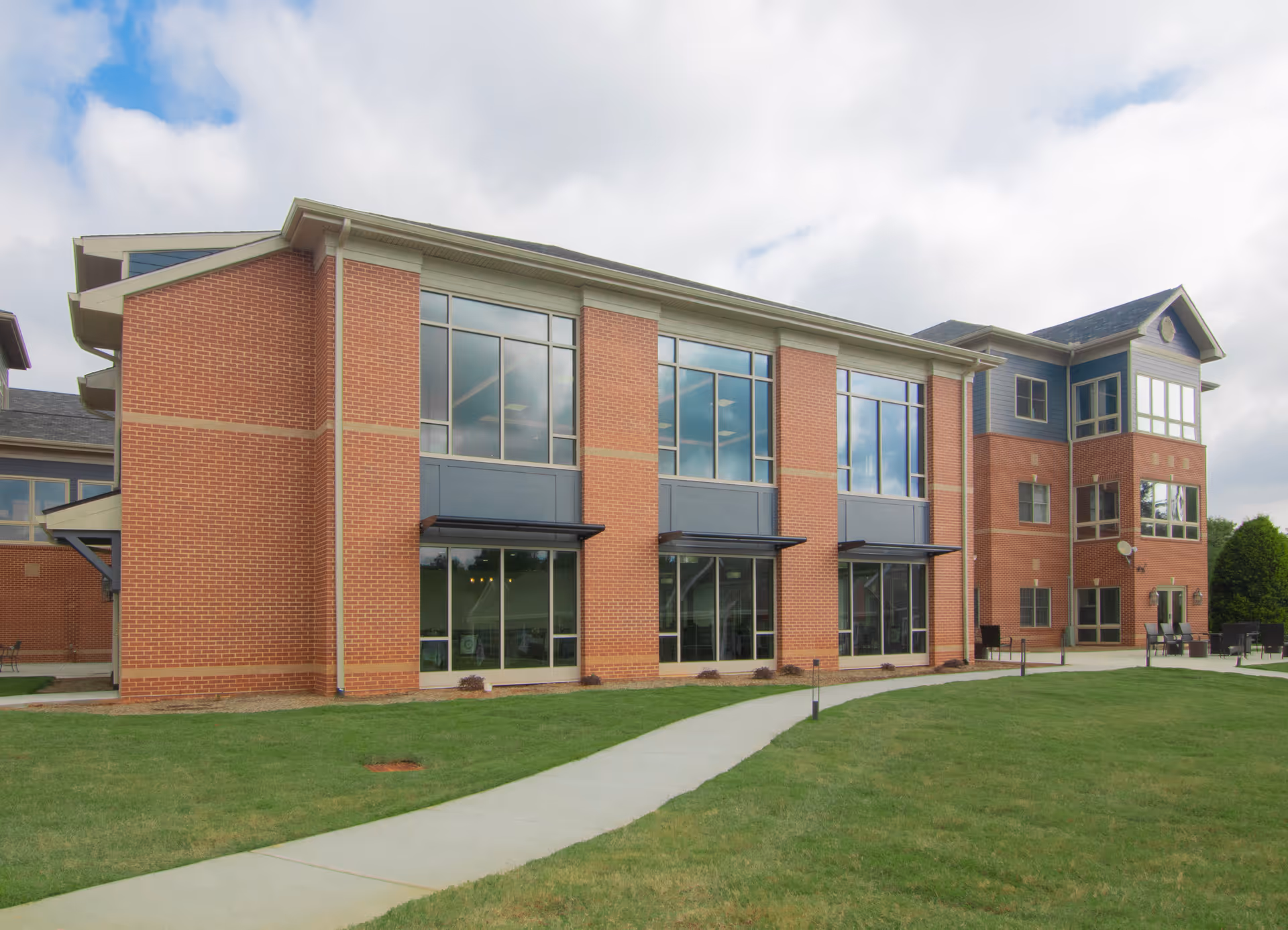 Exterior view of a multi-story brick building with large windows and a paved walkway leading to the entrance, surrounded by a well-maintained lawn under a partly cloudy sky.