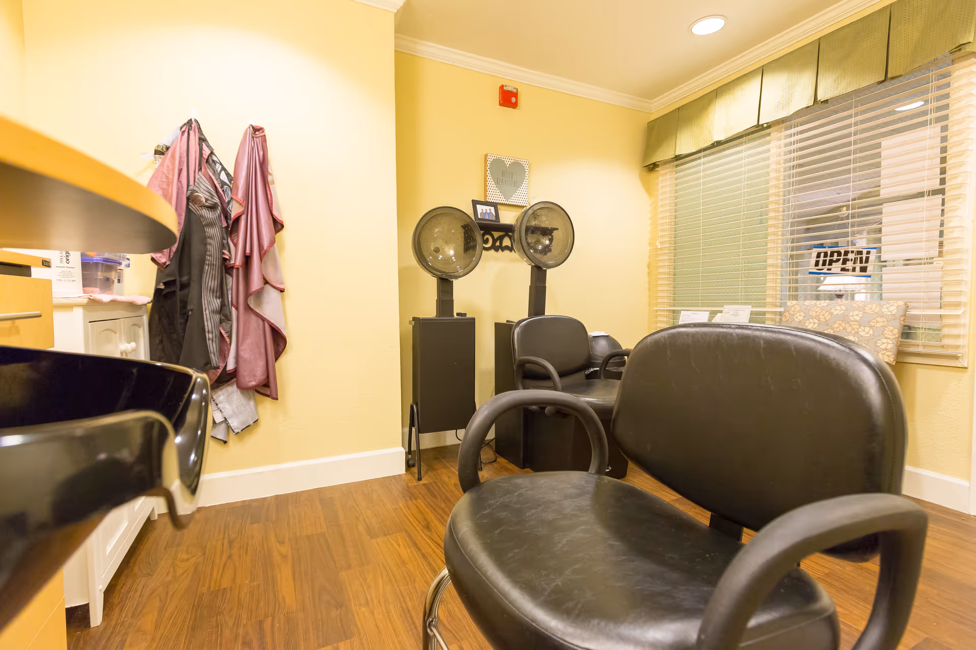Interior of a hair salon area with black salon chairs, hair dryers, and a window with blinds and a green valance. Several capes hang on hooks on a yellow wall, and the floor is wooden.