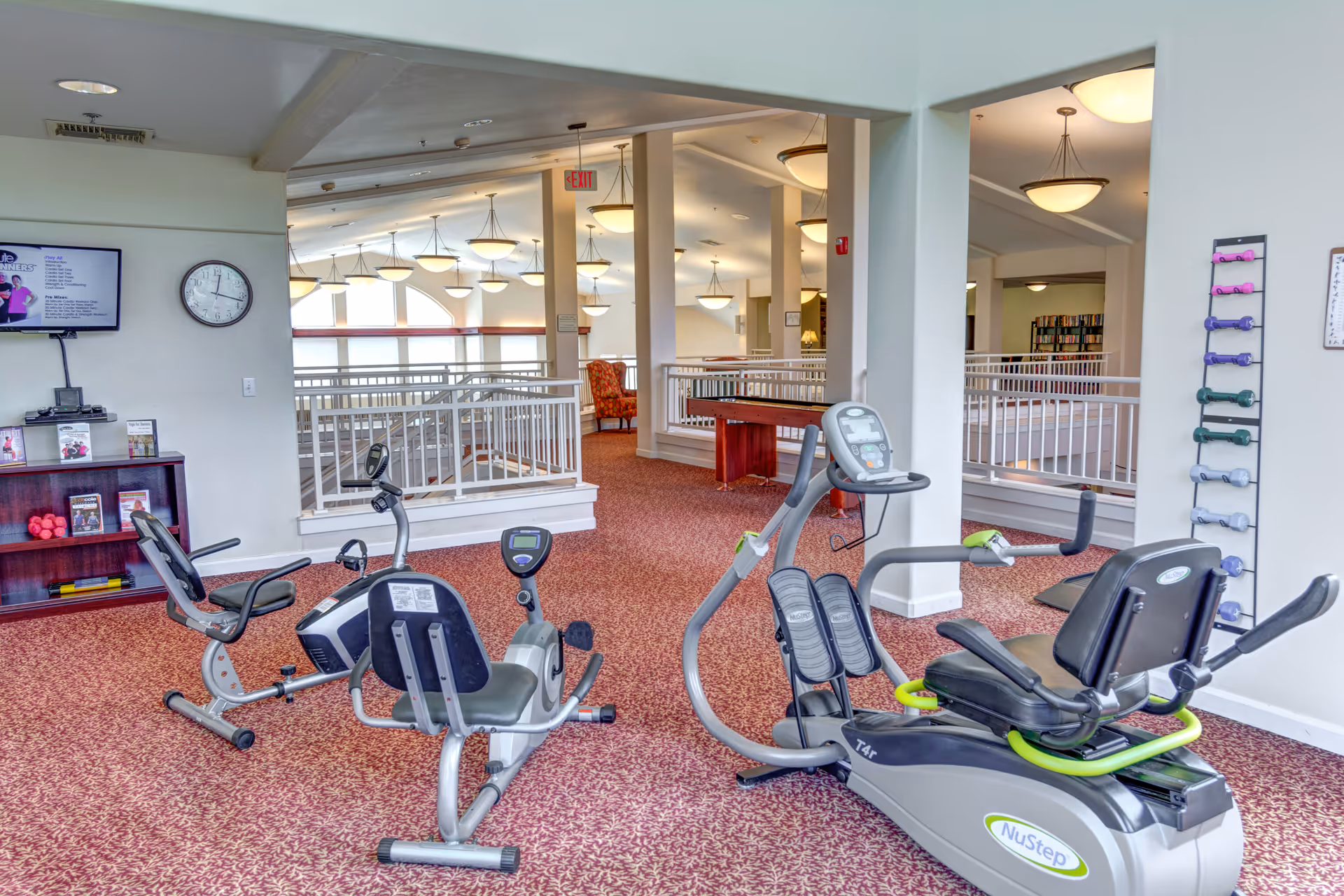An indoor exercise area in Hawks Ridge Assisted Living featuring two recumbent exercise bikes and a NuStep machine on a red patterned carpet. The room has white walls, multiple ceiling lights, a clock, a TV mounted on the wall, a rack with colorful dumbbells, and a bookshelf with exercise-related materials. The space overlooks a railing and an adjacent seating area with an orange armchair.