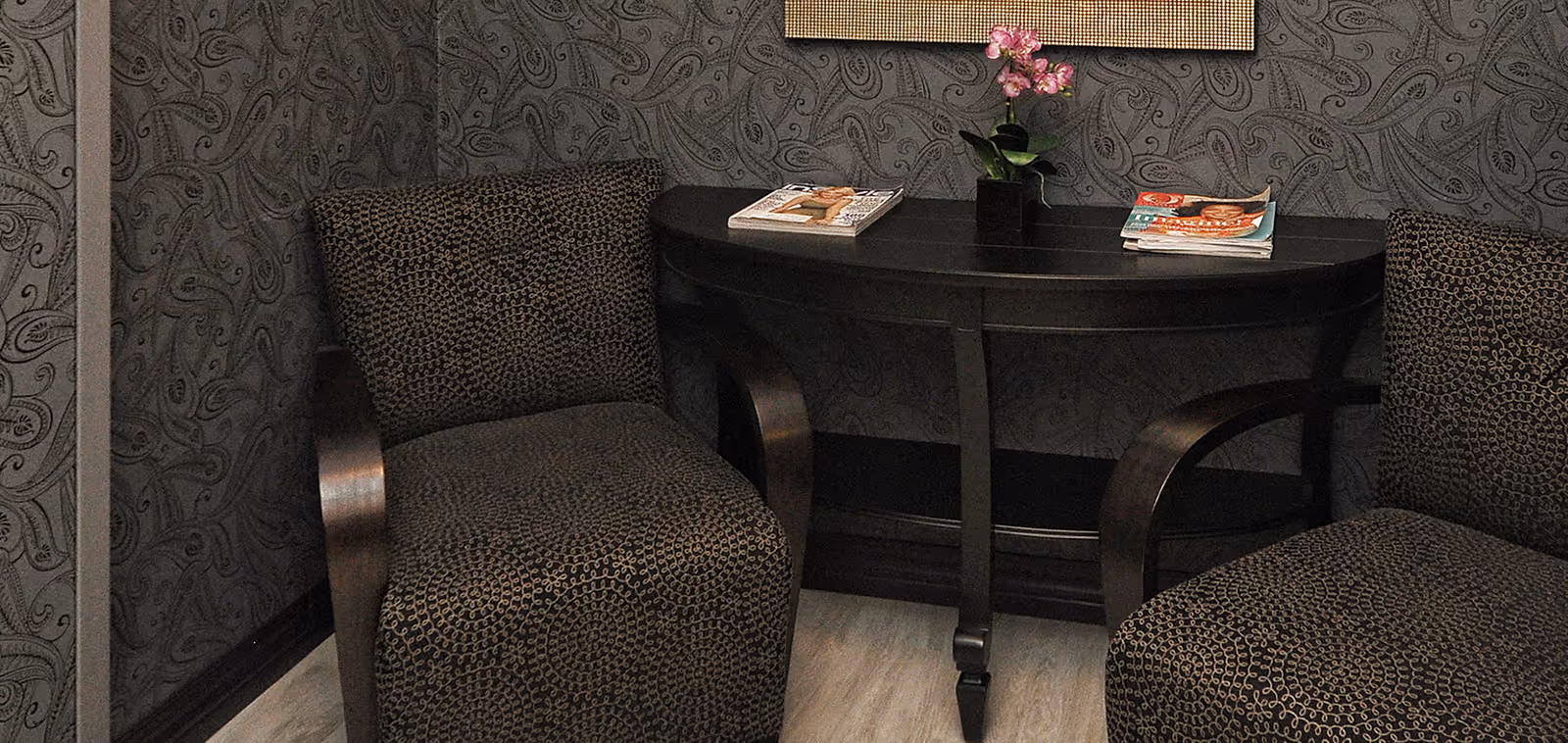 Two patterned armchairs positioned on either side of a black console table with curved legs. On the table are two stacks of magazines and a small potted plant with pink flowers. The background features a dark, intricately patterned wallpaper.