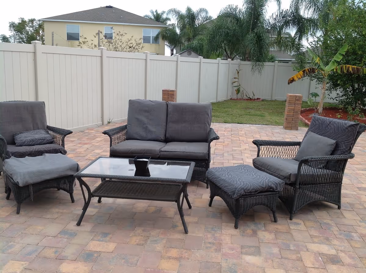 Outdoor patio area with a set of dark wicker furniture including a loveseat, two armchairs with cushions, two ottomans, and a glass-top coffee table. The patio is paved with brick tiles and enclosed by a white fence. There are some palm trees and other plants in the background along with a yellow two-story house visible over the fence.
