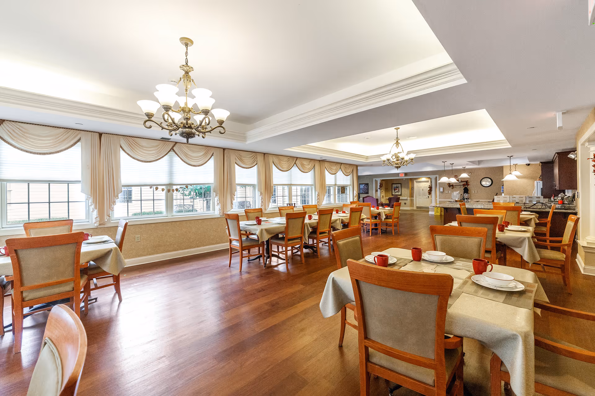Bright formal dining room with multiple set tables and chairs, chandeliers, large draped windows, and a serving area in the background.