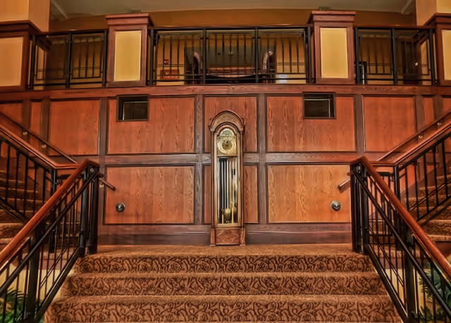 Carpeted staircase with ornate patterned carpet leading up to a landing featuring a wooden grandfather clock set into a wood-paneled wall. The staircase has black metal railings with wooden handrails, and the upper level has a railing with vertical bars and decorative panels.