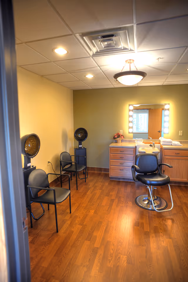 Interior salon room with a styling chair, hood hair dryers, a lighted vanity mirror, and waiting chairs on wood flooring.