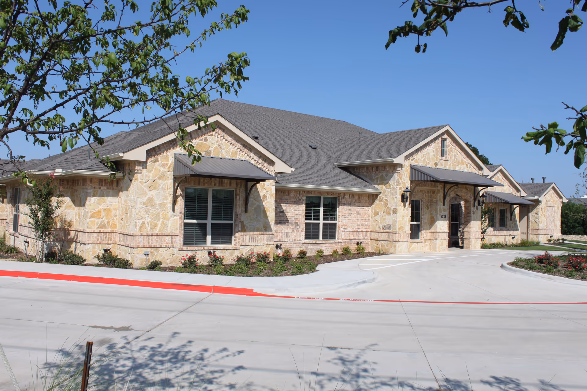 Exterior view of a single-story stone and brick building with a gray roof under a clear blue sky. The building has multiple windows with awnings and a curved driveway in front, surrounded by landscaped bushes and small trees.
